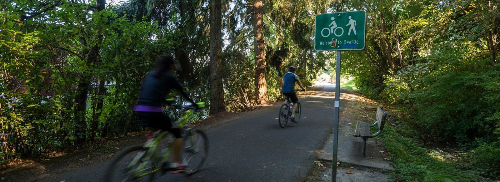 Two cyclists on a paved path through trees, beside a trail sign.
