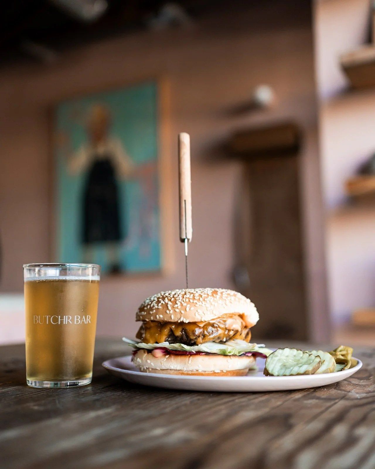 A burger on a plate beside a glass of beer on a wooden table, with a painting on the pink wall behind