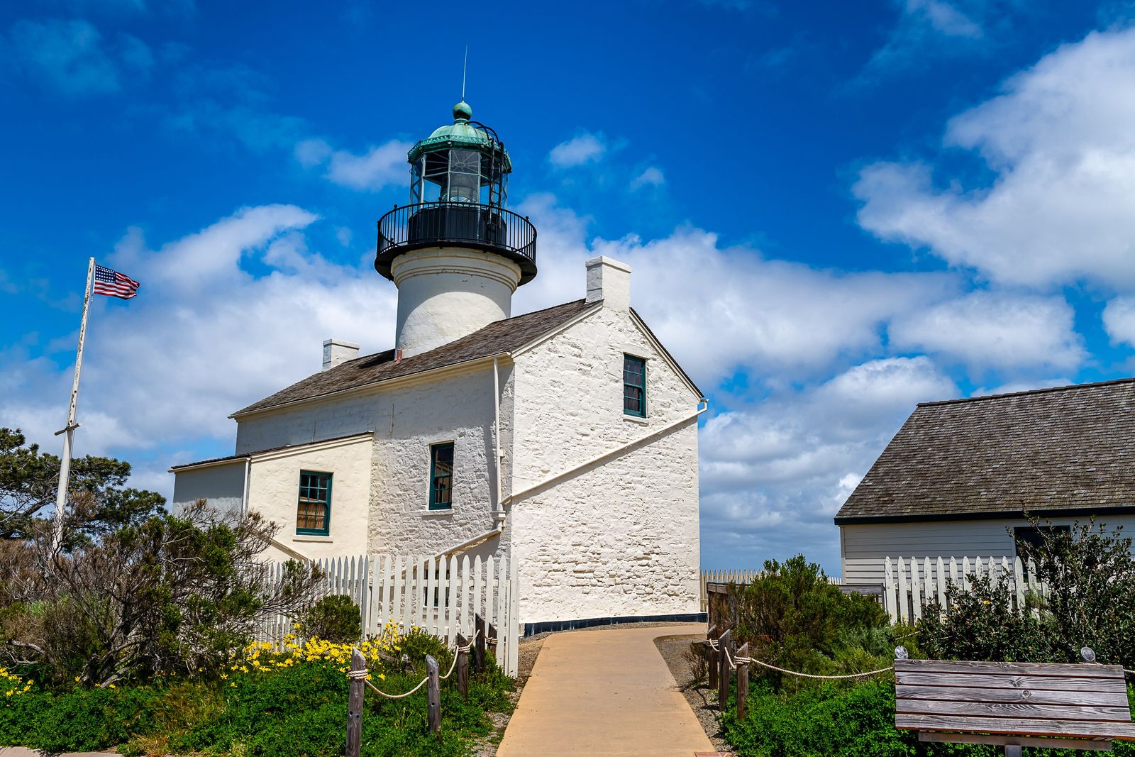 The white old lighthouse with a dark lantern room on a hilltop under a blue sky with clouds.