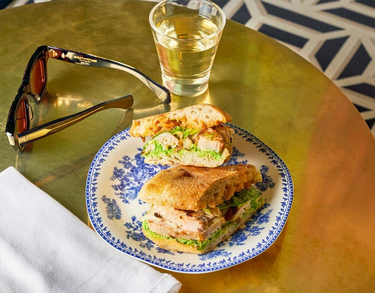 A sandwich halved on a blue-and-white plate beside a glass and sunglasses on a gold table.