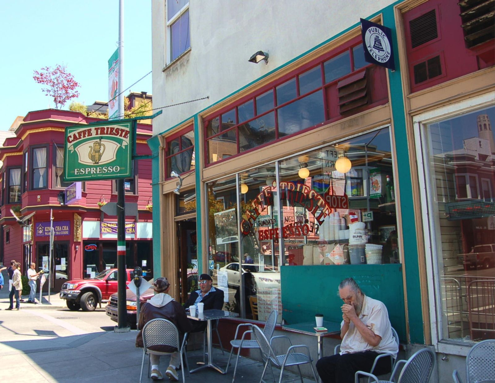 A cafe corner with a green awning and outdoor seating under a street sign, with a customer at a table.