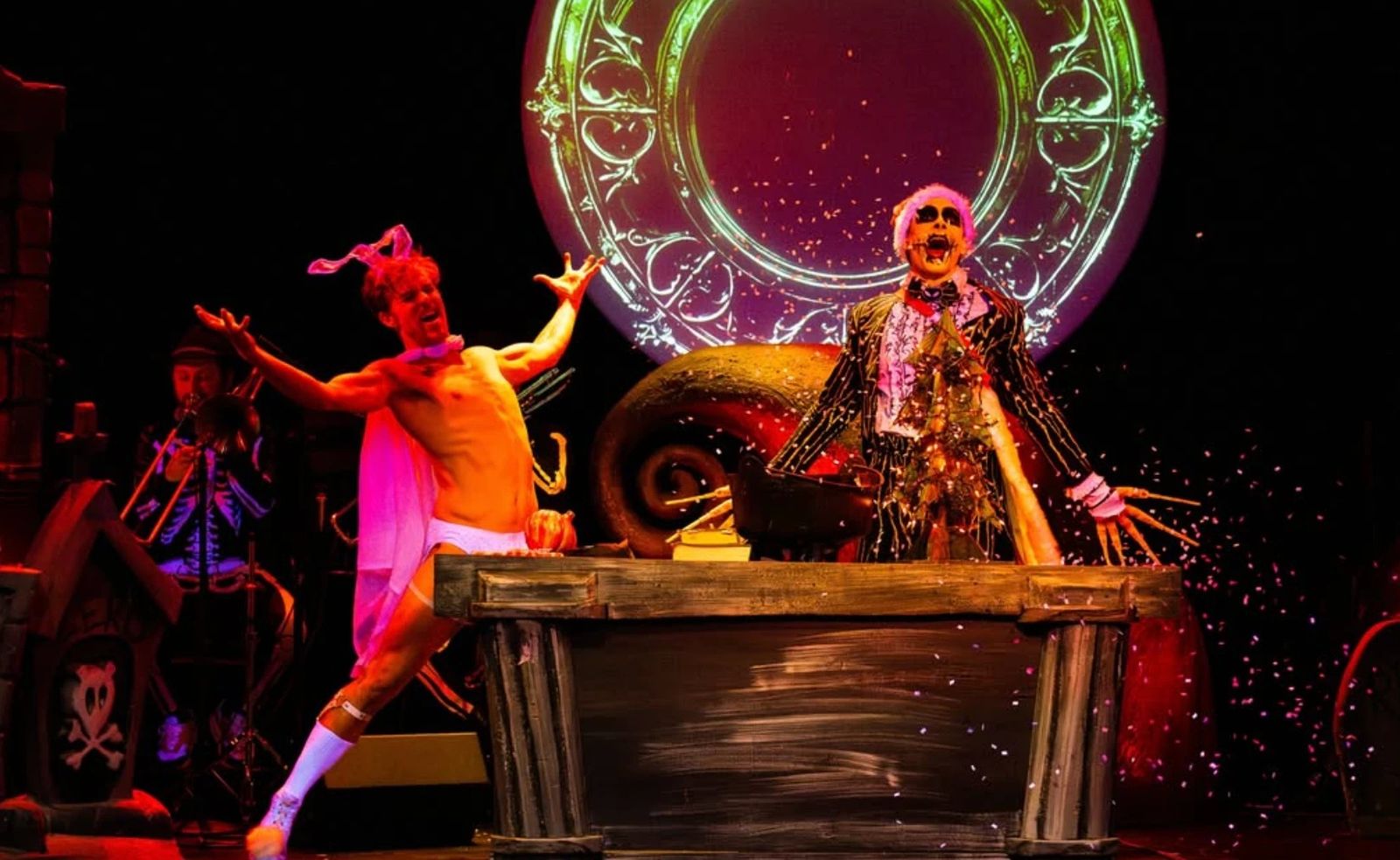 Two performers on a dark stage, one in a pink cape and bunny ears and one in a skeleton-face costume and pinstripe suit, with confetti in the air.