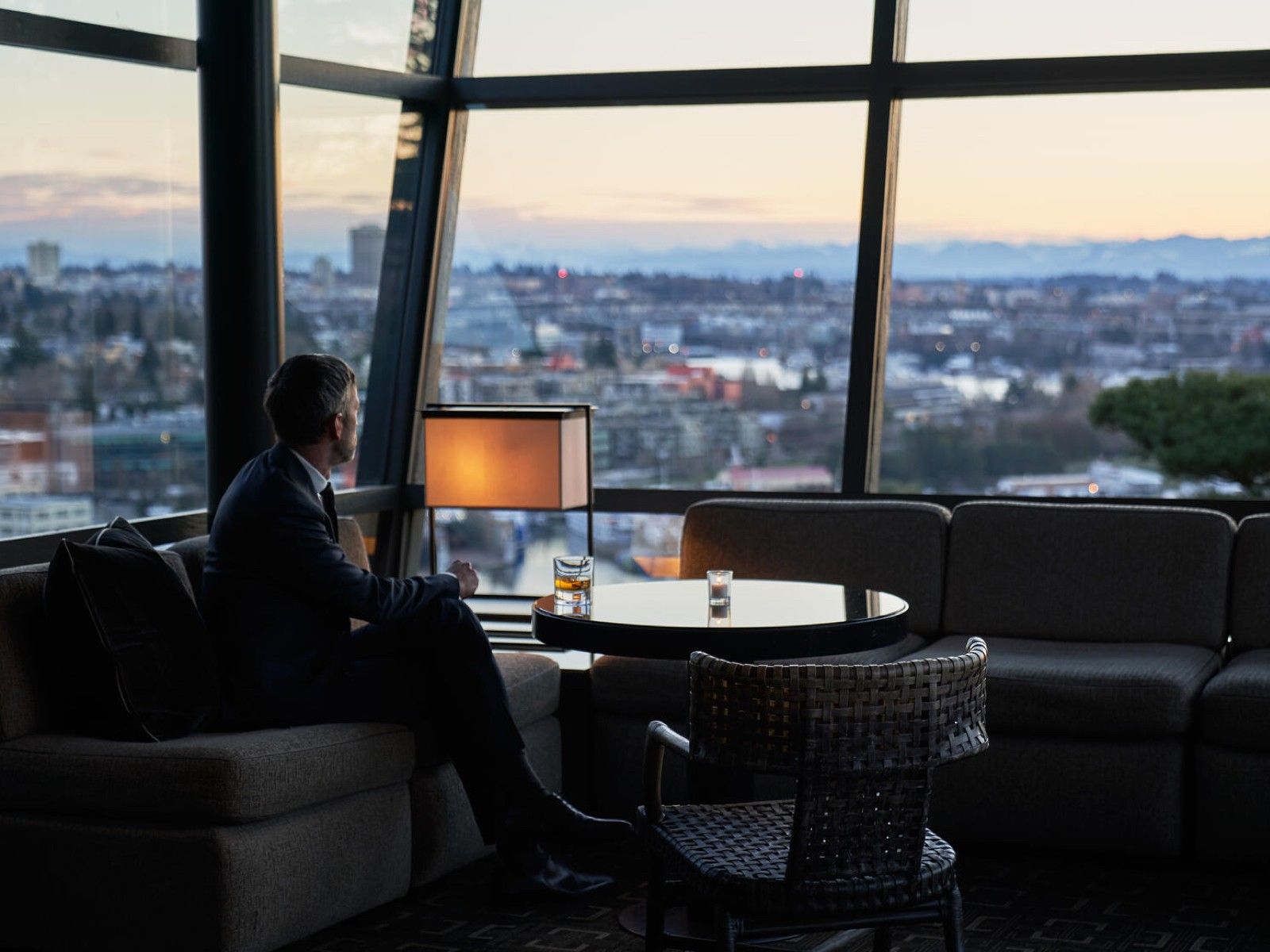 A lounge with low seating and tall windows looking out over the city at dusk, with one person seated.