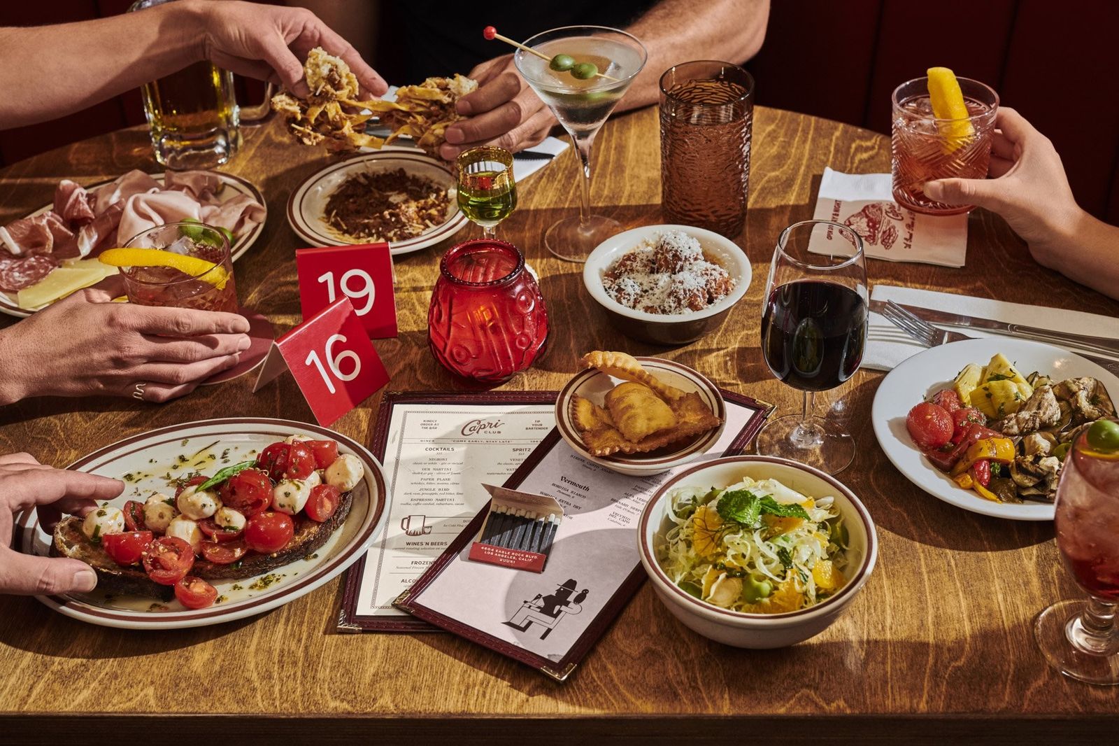 A table of shared plates, glasses of wine and cocktails, with several hands reaching in.