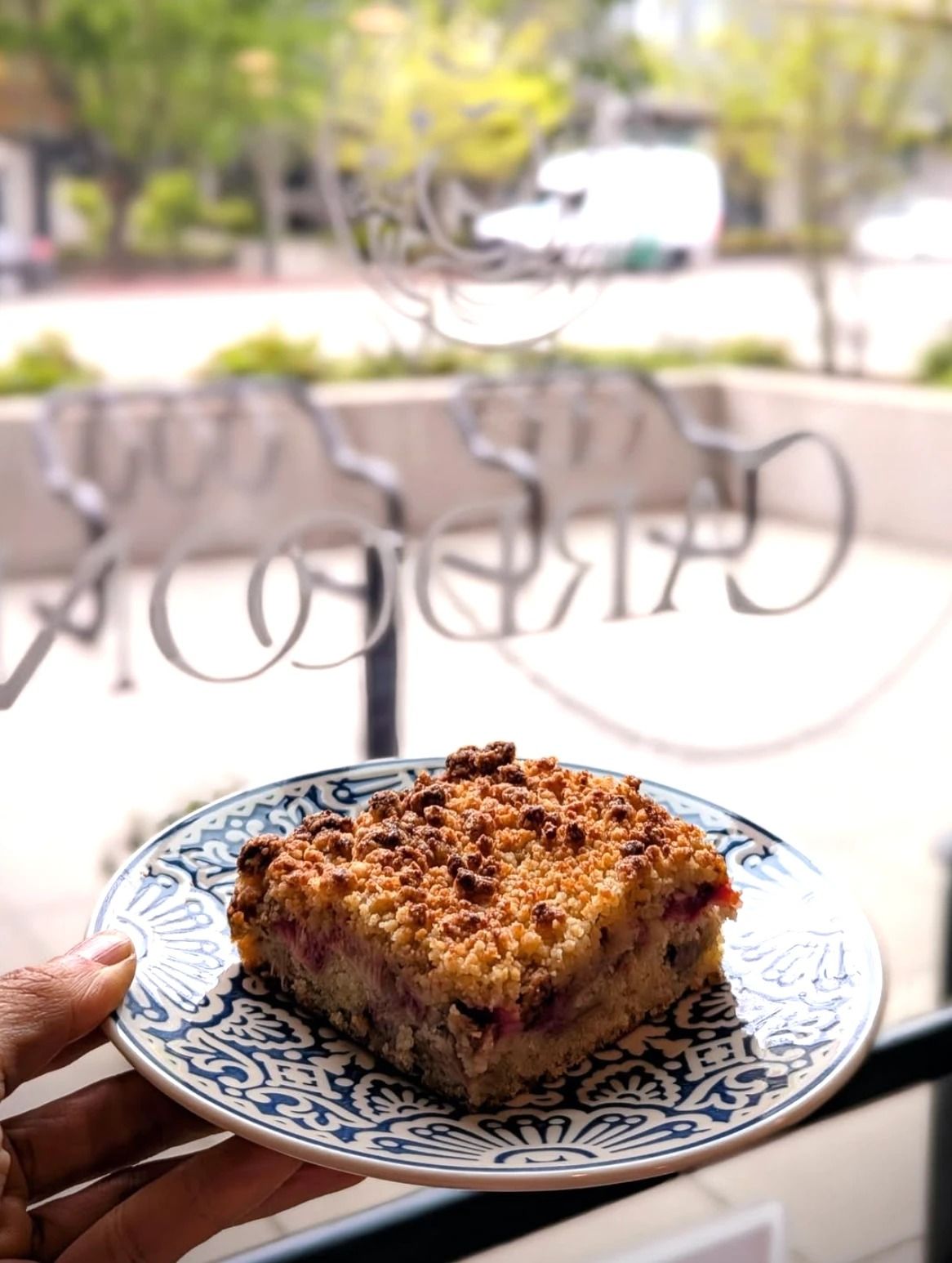A slice of crumb-topped fruit cake on a blue-and-white plate held up in front of a window.