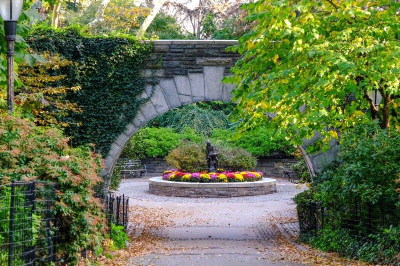 A stone archway over a flower bed with green foliage in Carl Schurz Park