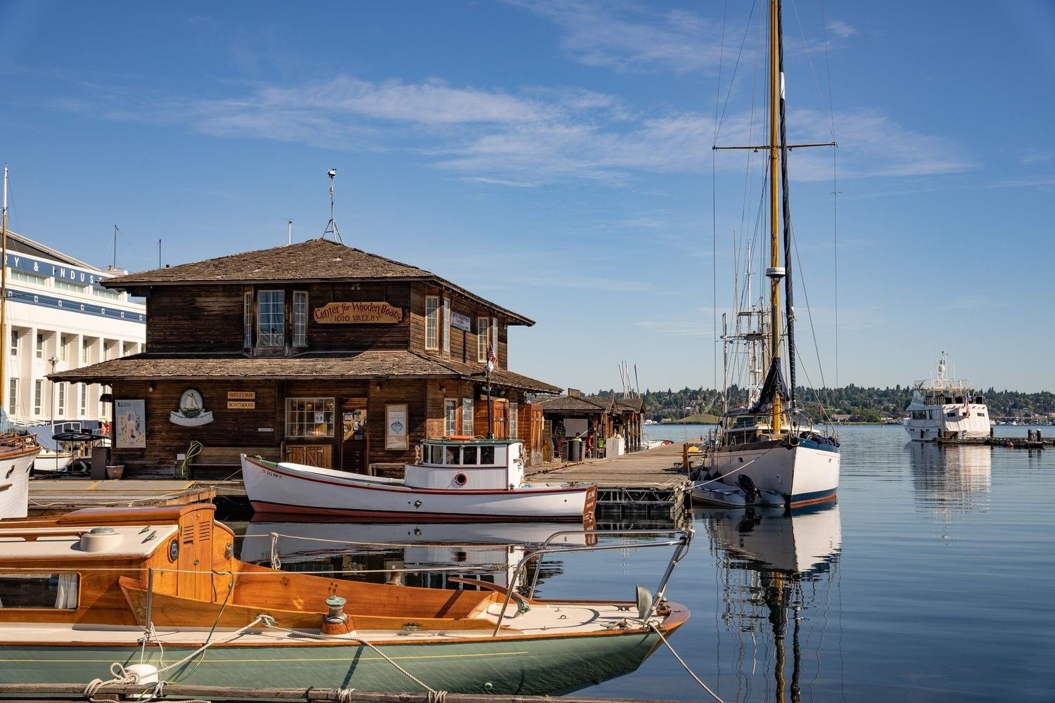 A wooden boathouse and moored sailboats on calm water under a blue sky.