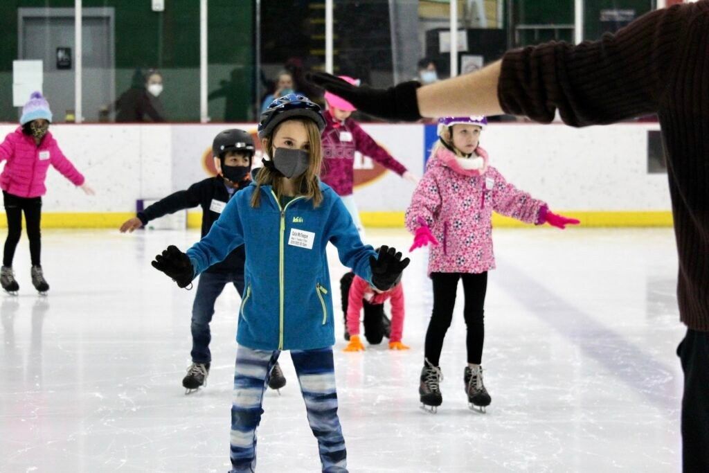 A child in a blue jacket skating on an indoor rink with other skaters behind.