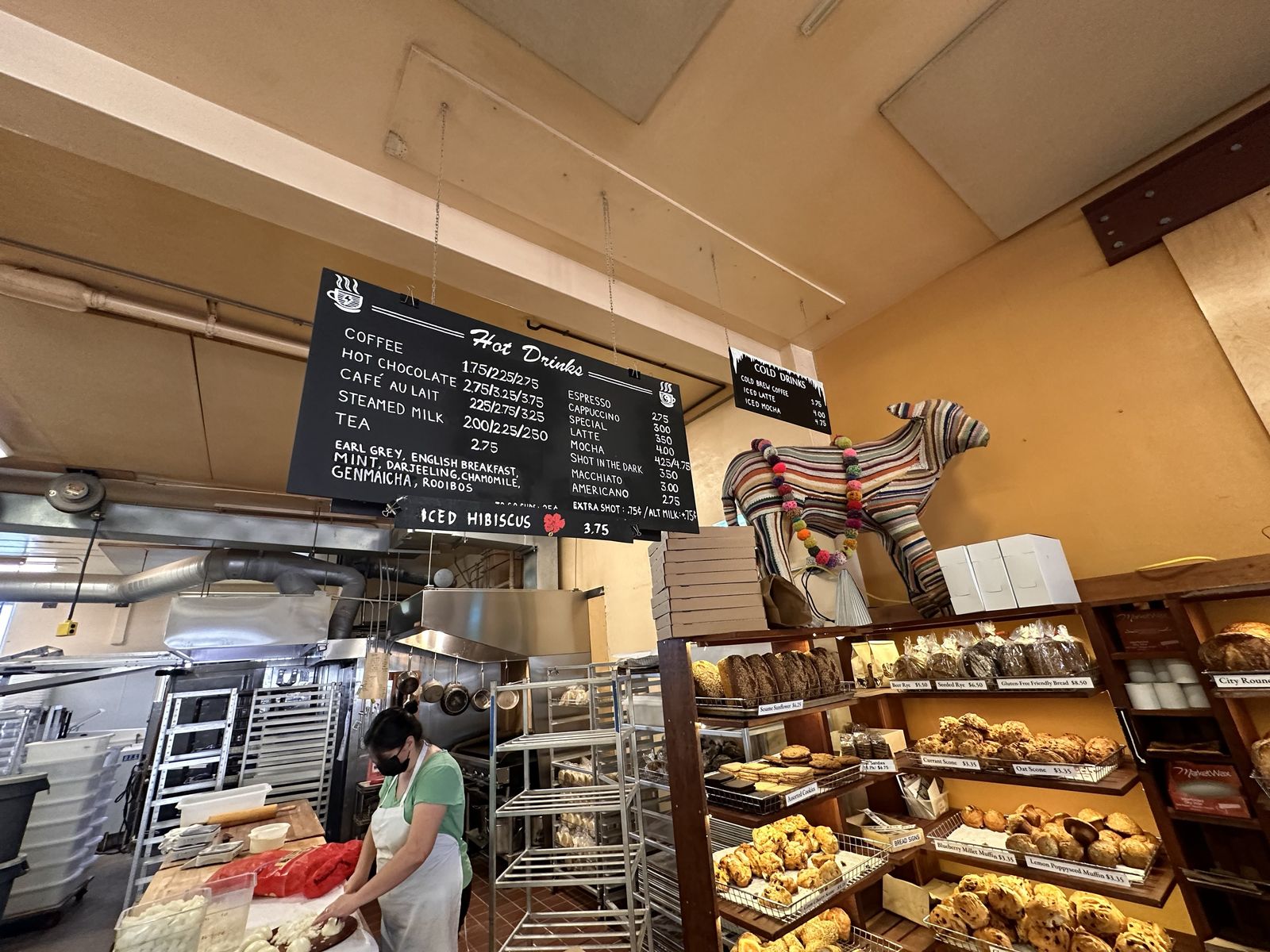 A busy bakery counter with shelves of loaves, a chalkboard menu and a worker behind the counter.