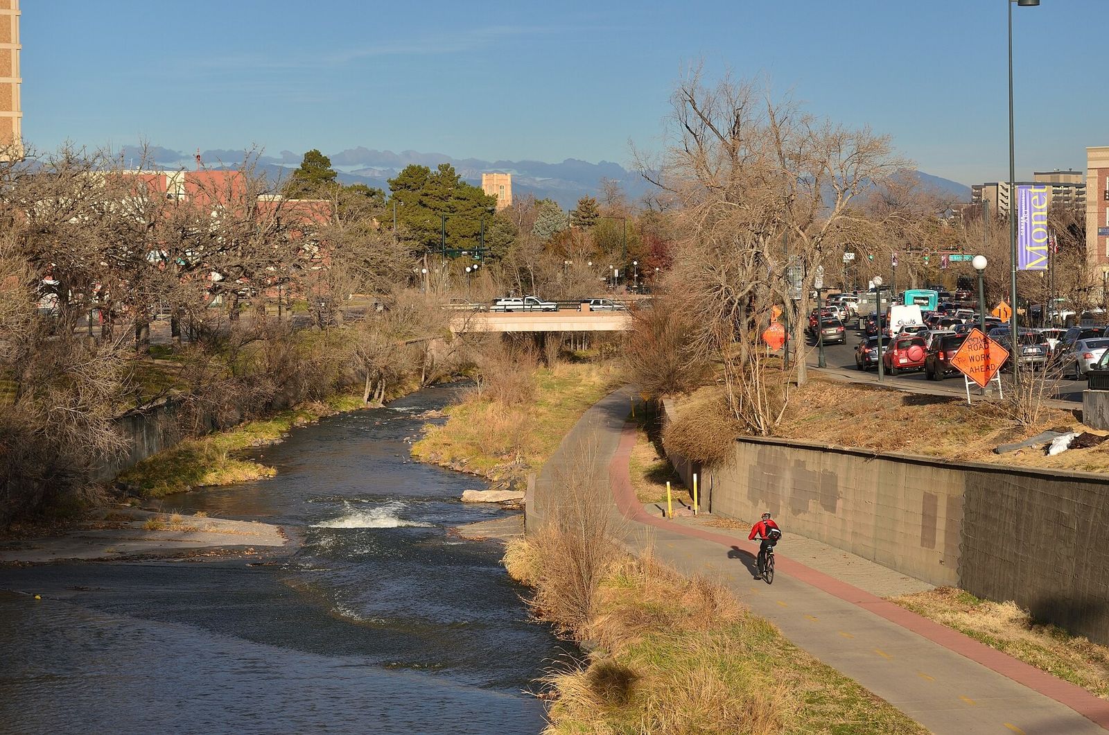 A creek running between a paved trail and banks of bare trees, with a cyclist and mountains in the distance.