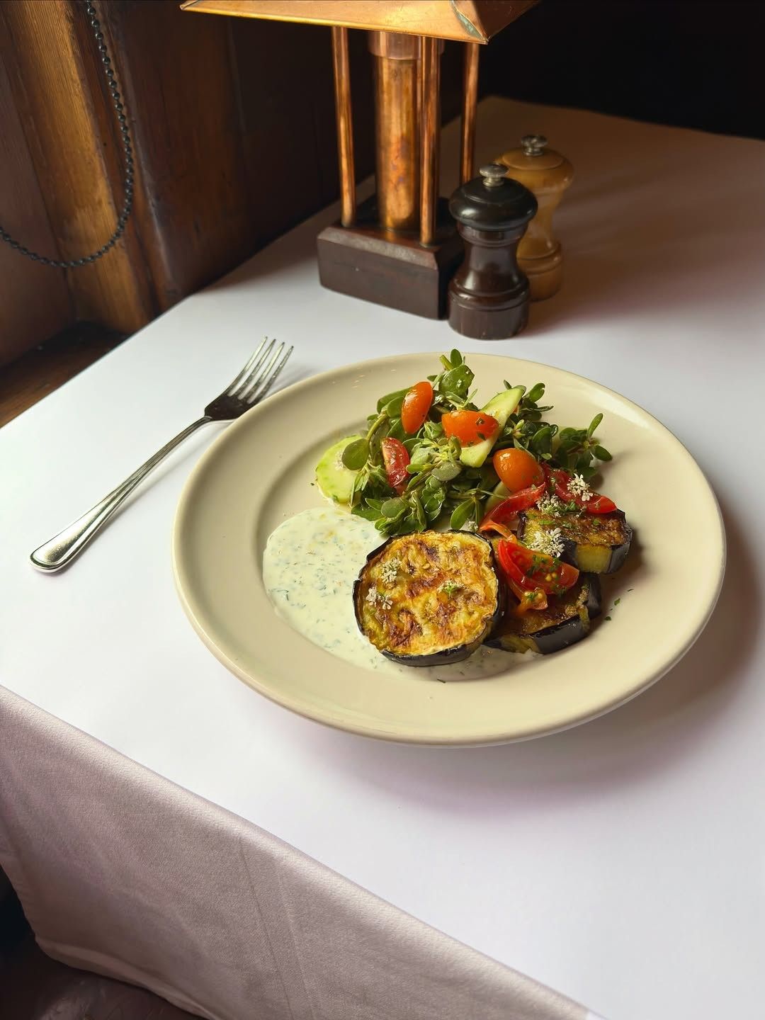 A white-clothed table with a plate of salad and grilled eggplant, a fork and a pepper mill.