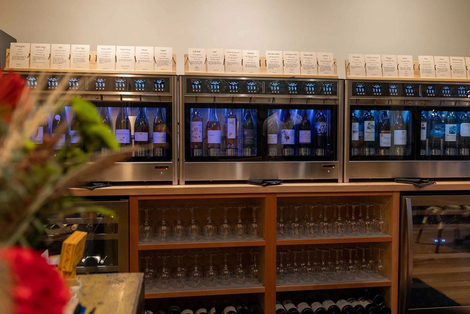 A wall of wine dispensing machines above a wooden counter and shelves of glasses.