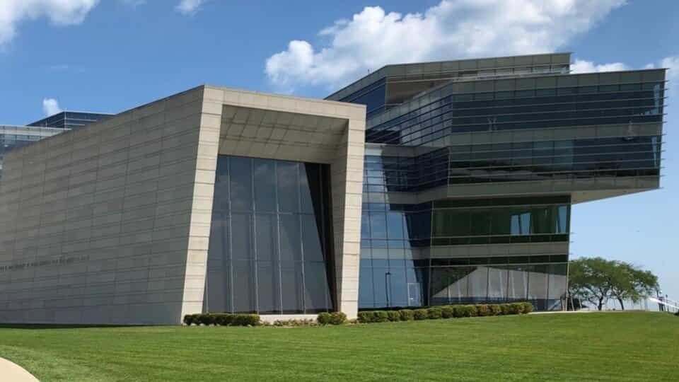 A modern building of gray stone and dark glass cantilevered over a lawn under a blue sky with clouds.