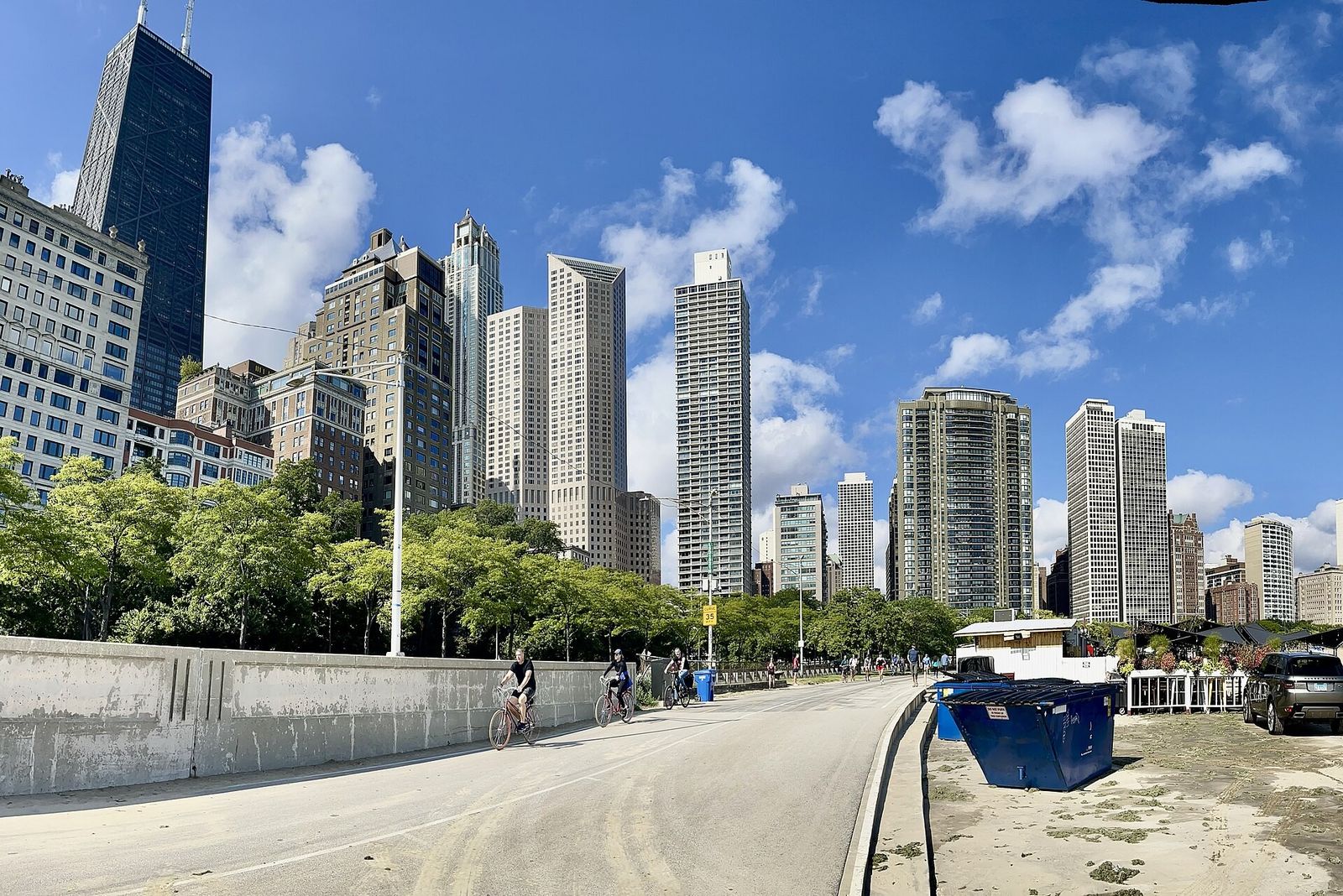 A paved lakefront path beside a low wall with a beach, high-rise towers and a blue sky with clouds.