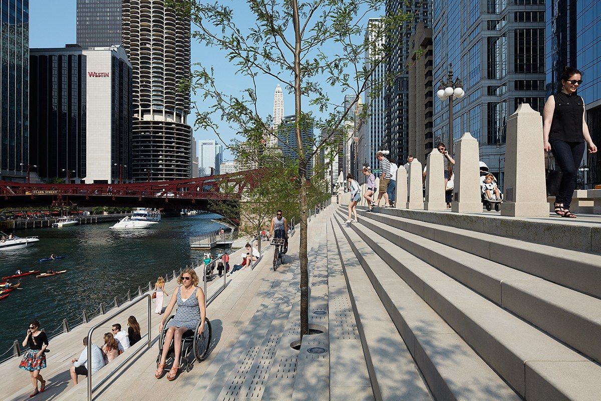 A wide stepped riverwalk beside the river with walkers, trees and downtown towers on both banks.