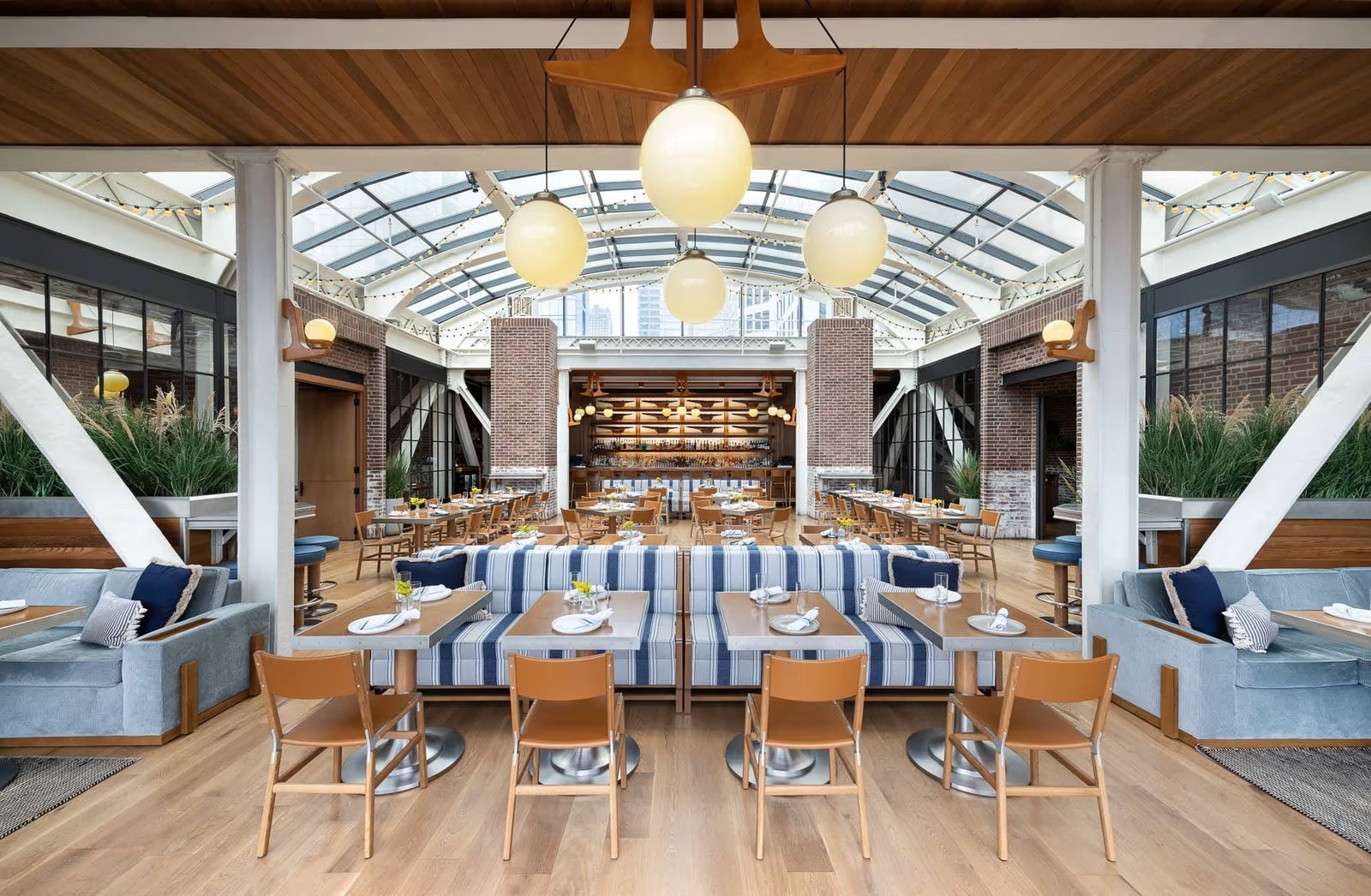 A glass-roofed dining room with striped banquettes, wooden tables and hanging globe lights.
