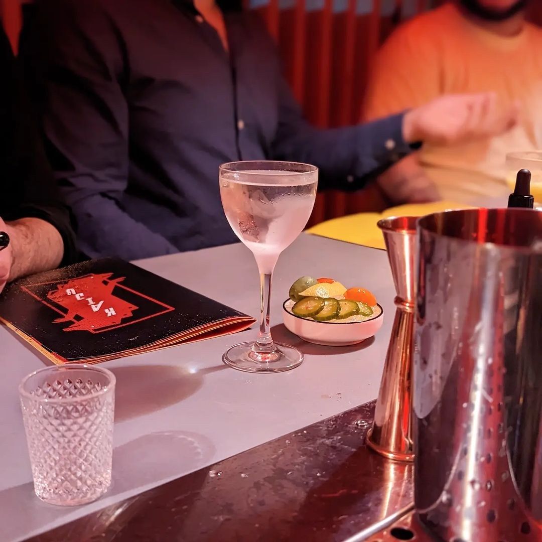 A cocktail glass, shaker and menu on a pink bar top with a guest in the background