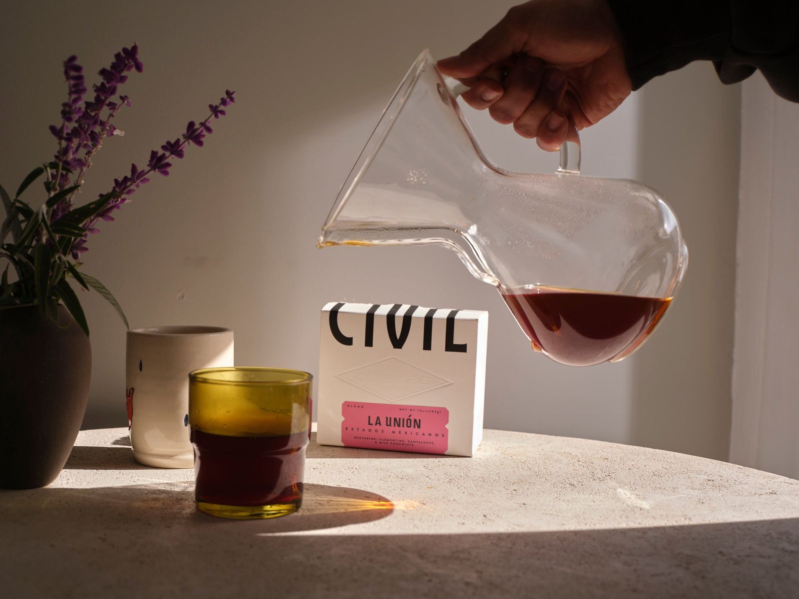 A hand pouring coffee from a glass server beside a mug and a cup, with a CIVIL sign on the table.