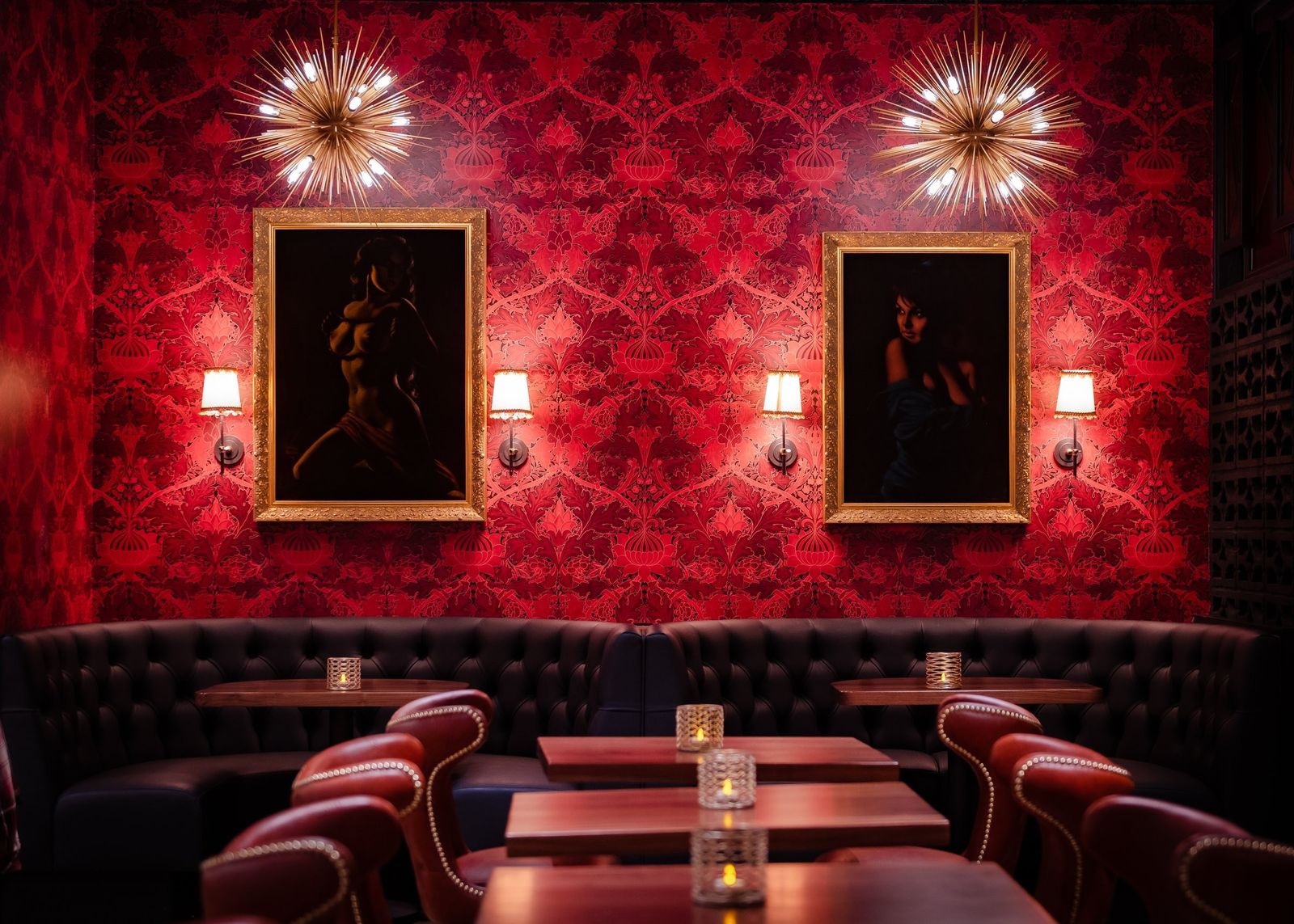 A lounge with a red velvet banquette, round tables and framed paintings on a patterned red wall lit by wall sconces.