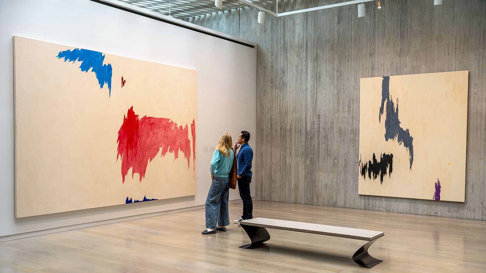 A gallery with concrete walls and a wooden floor, with large abstract paintings and two visitors beside a bench.
