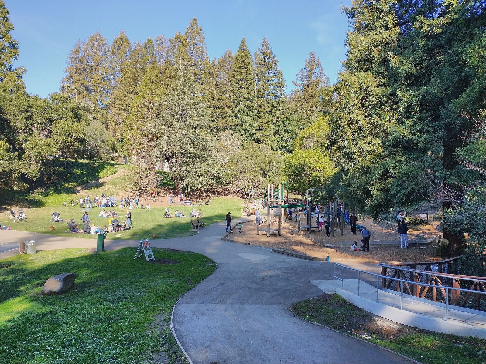 A paved path curving through a wooded park with a playground and visitors on the grass.