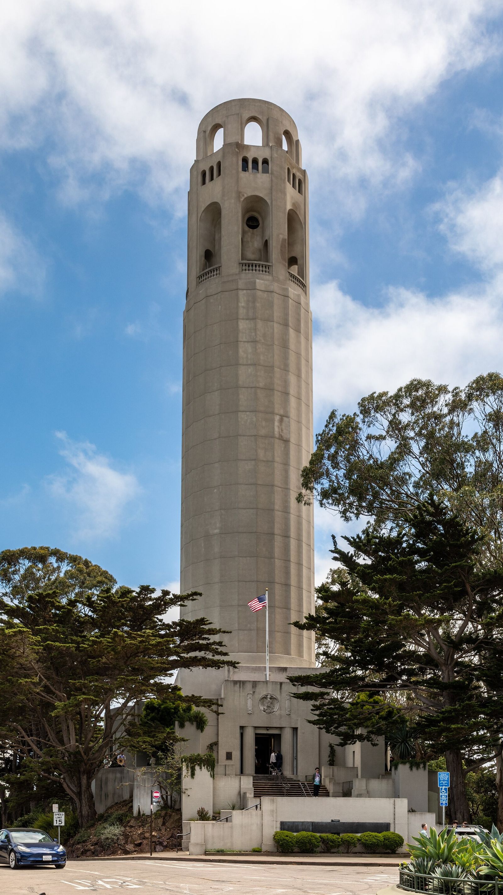 The tall fluted concrete tower of Coit Tower rising above trees under a blue sky, seen from its base.