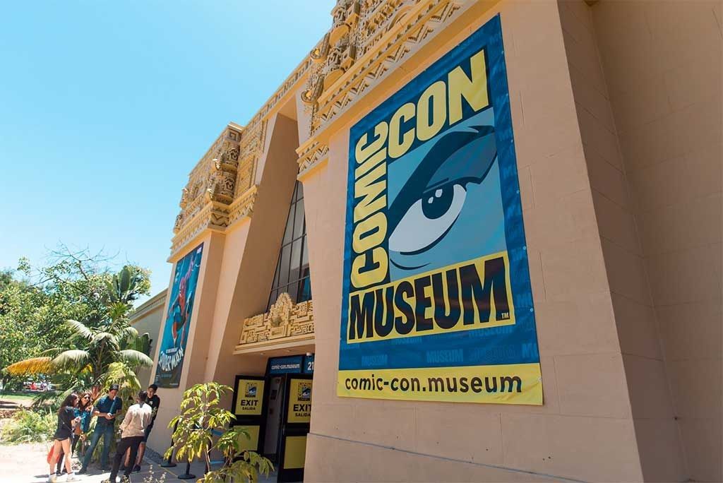 The sandy-colored building facade with a large Comic-Con Museum banner, palms and visitors below.