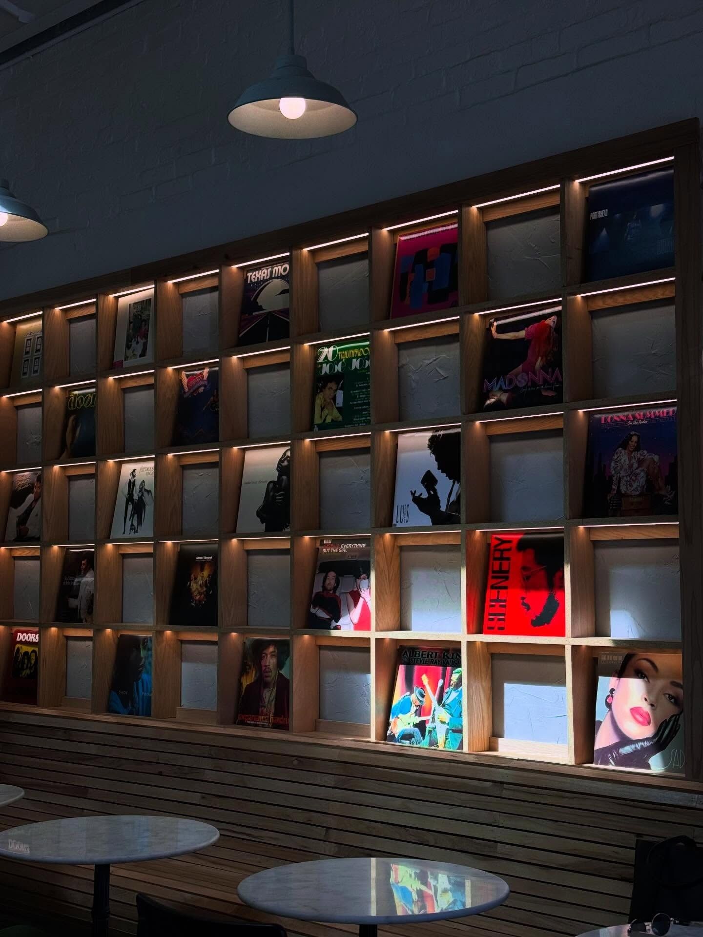 A wall of shelves holding framed vinyl record sleeves, lit from within, above two small round cafe tables.