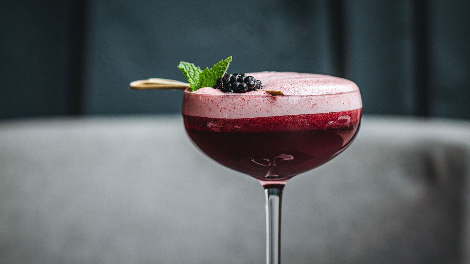 A coupe glass of a purple frothy cocktail garnished with mint and a blackberry on a pick.