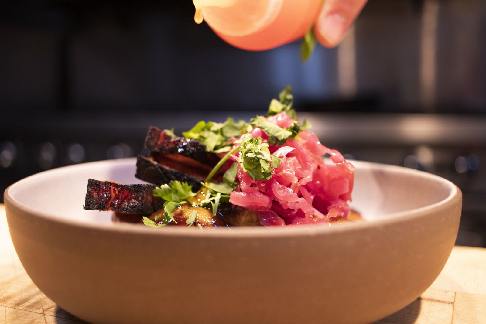 A shallow clay bowl of braised meat with greens and pink pickled onion, finished with a drizzle from a spoon.