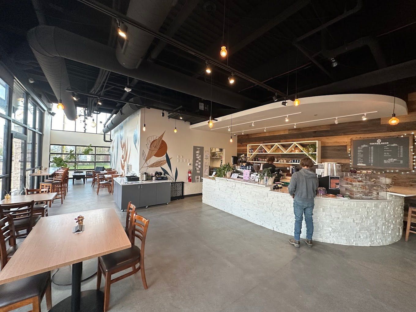 A spacious cafe with a white tiled counter, a customer ordering, wooden tables and high windows.