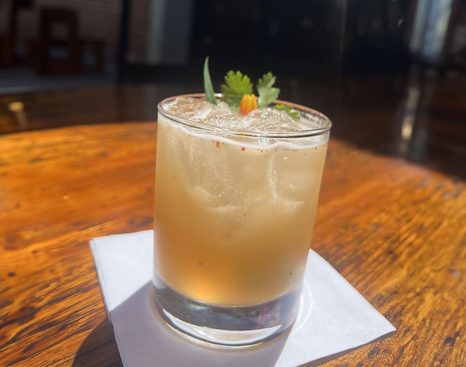 A cocktail with a citrus peel and a herb sprig in a short glass on a worn wooden bar top.
