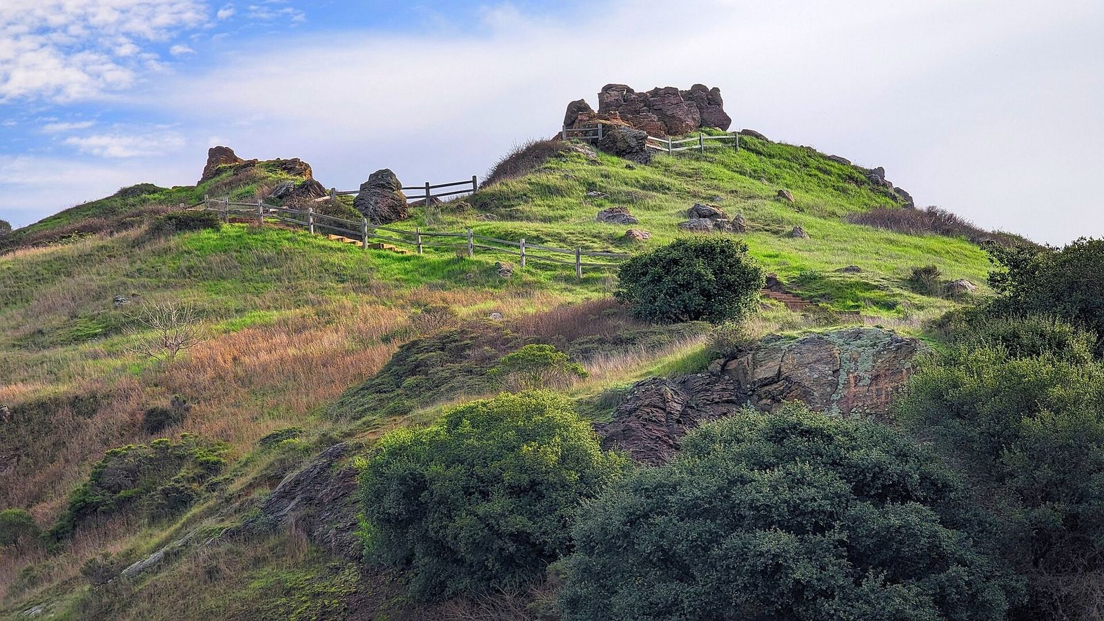 A grassy hillside with scrub and rocky outcrops under a blue sky with clouds.