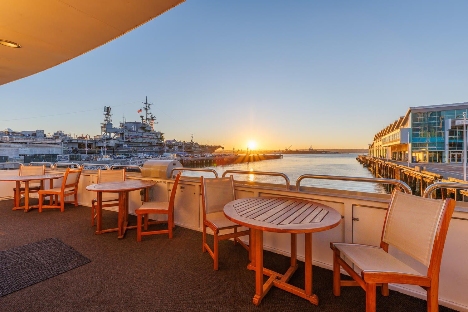 Wooden tables and chairs on a boat deck with a sunset over the harbor and a navy ship beyond.