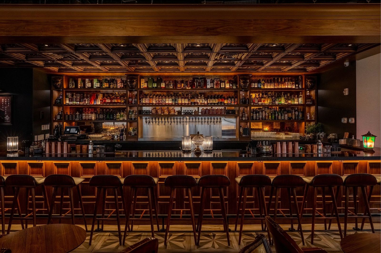 A long bar with a lit shelf wall of bottles above a row of stools in a warmly lit room.
