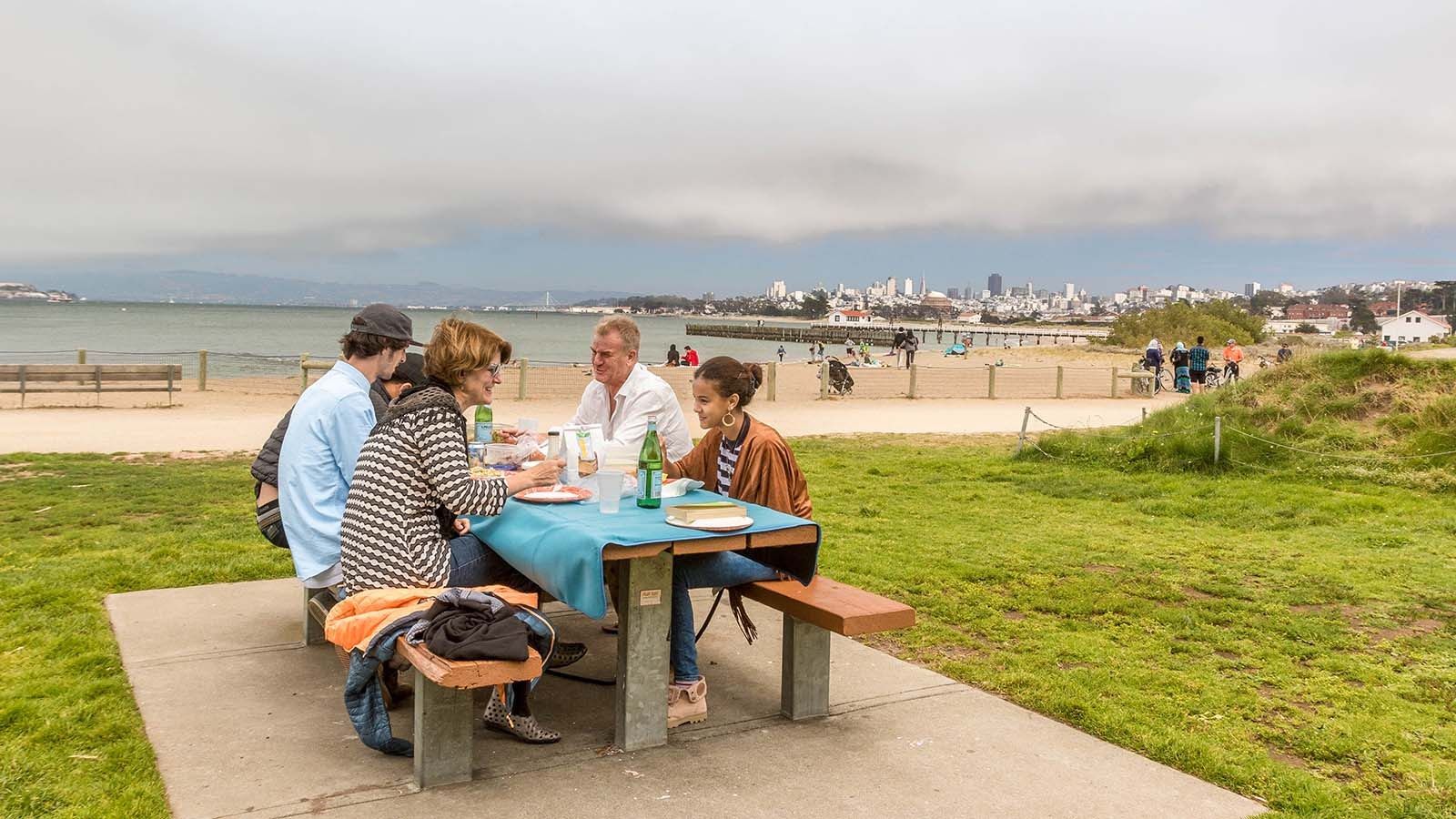 A group at a picnic table on grass by a beach, with a hazy bay and city skyline behind.