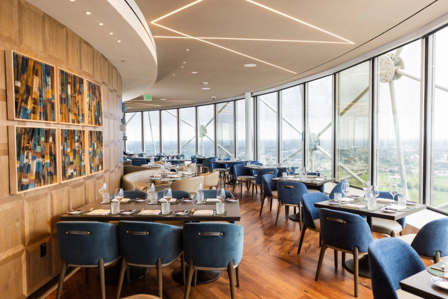 A curved dining room with floor-to-ceiling windows, blue chairs and framed art along a wood-paneled wall.
