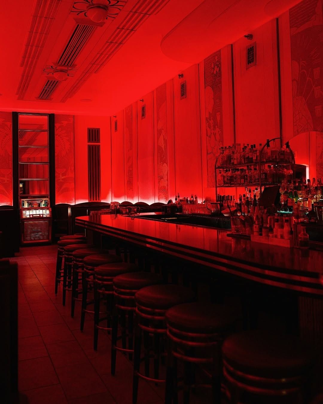 A long bar lit in deep red light, with stools along the counter and rows of bottles on the back wall.