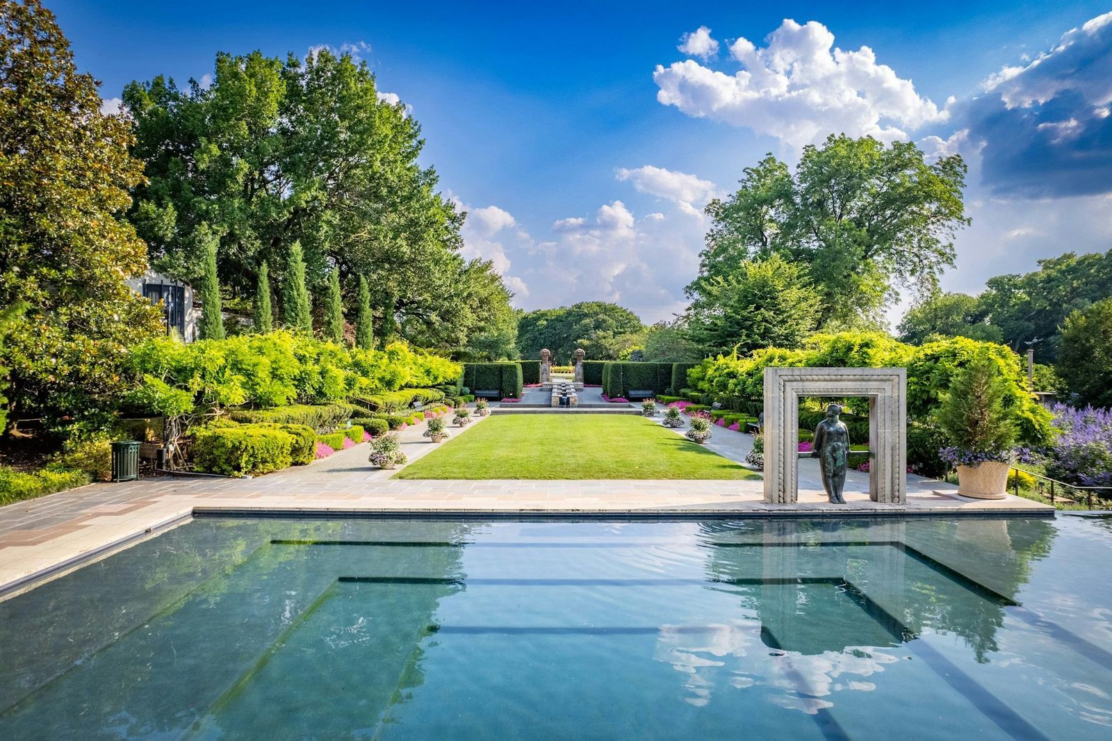 A long reflecting pool leading to a garden gate between rows of tall hedges under a blue sky.