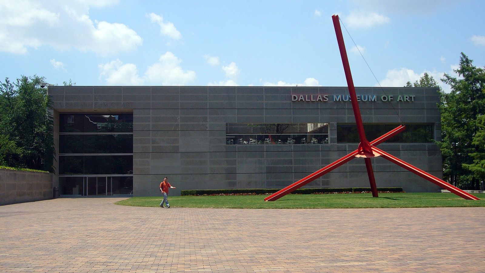 A gray museum building with a large red sculpture of angled beams in front, on a brick plaza.