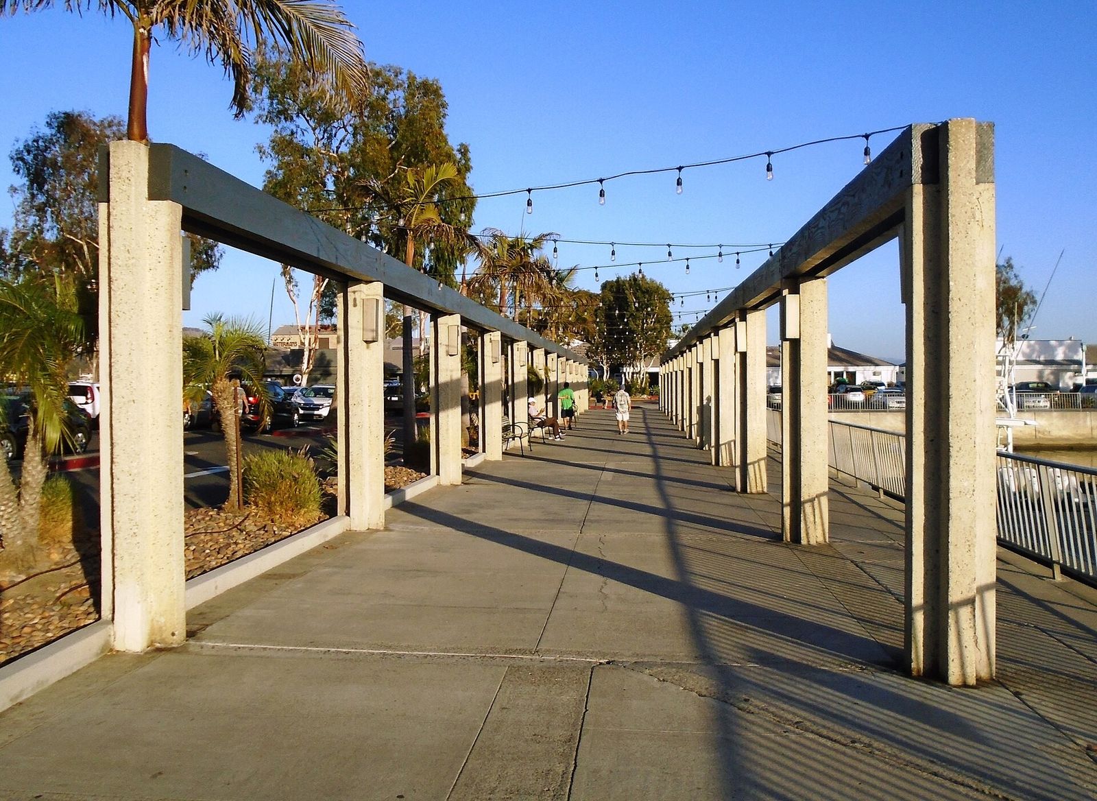 A shaded walkway under pergola columns with string lights, beside a railing and harbor.