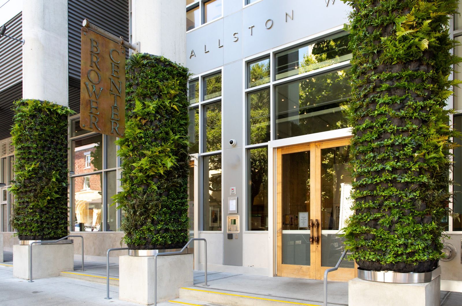 A glass-fronted building entrance with wooden doors and tall vertical walls of green plants on either side.
