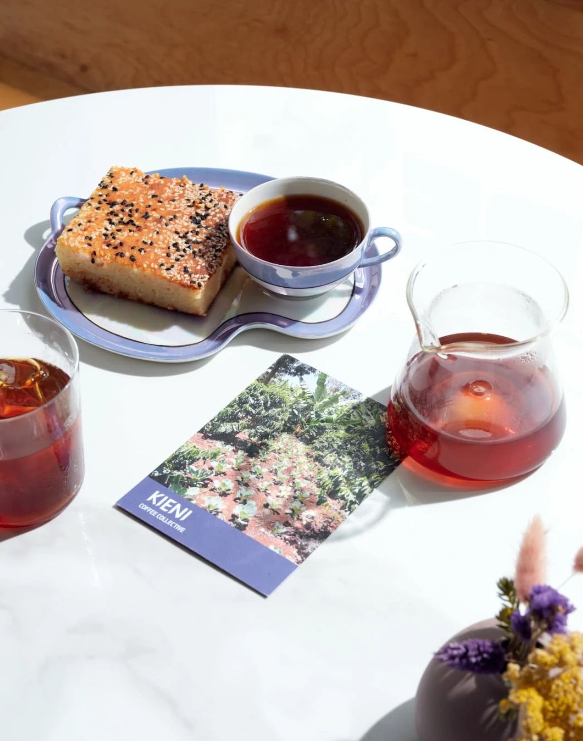 A slice of sesame-topped cake and a cup of black coffee on a lilac plate, with a carafe and a glass of coffee on a white table.