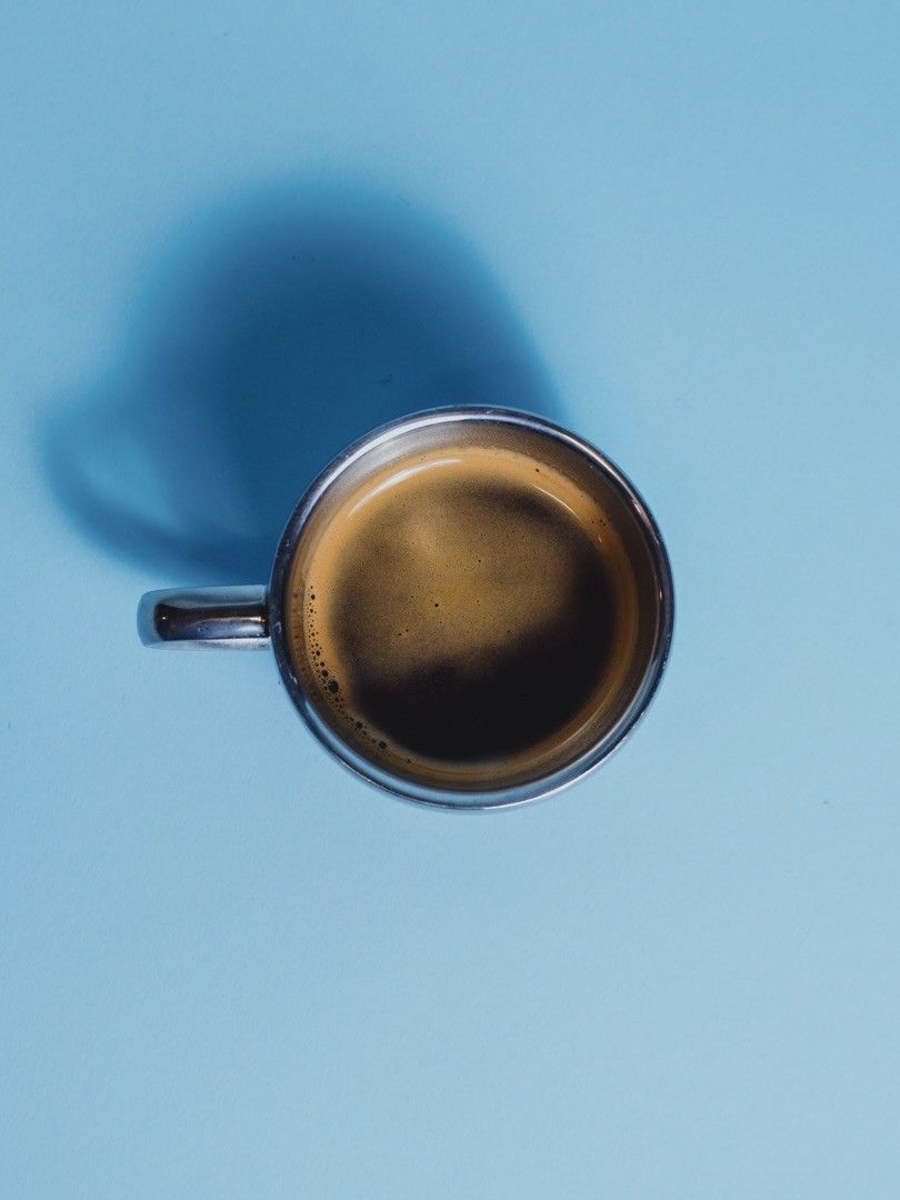 An espresso in a small glass cup seen from above on a light blue surface with a long shadow.