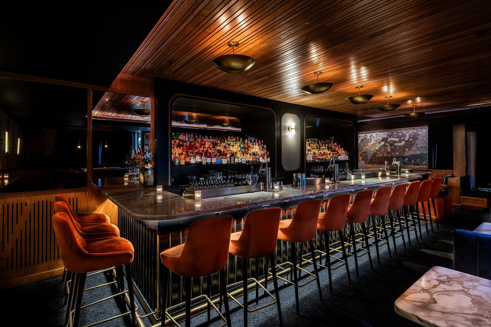 A dark bar with a long counter of orange leather stools, a wood-slat ceiling and a lit back bar.