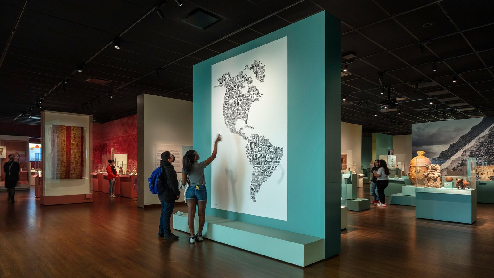 A dim gallery with a tall teal display of a map of the Americas and visitors reading the wall.