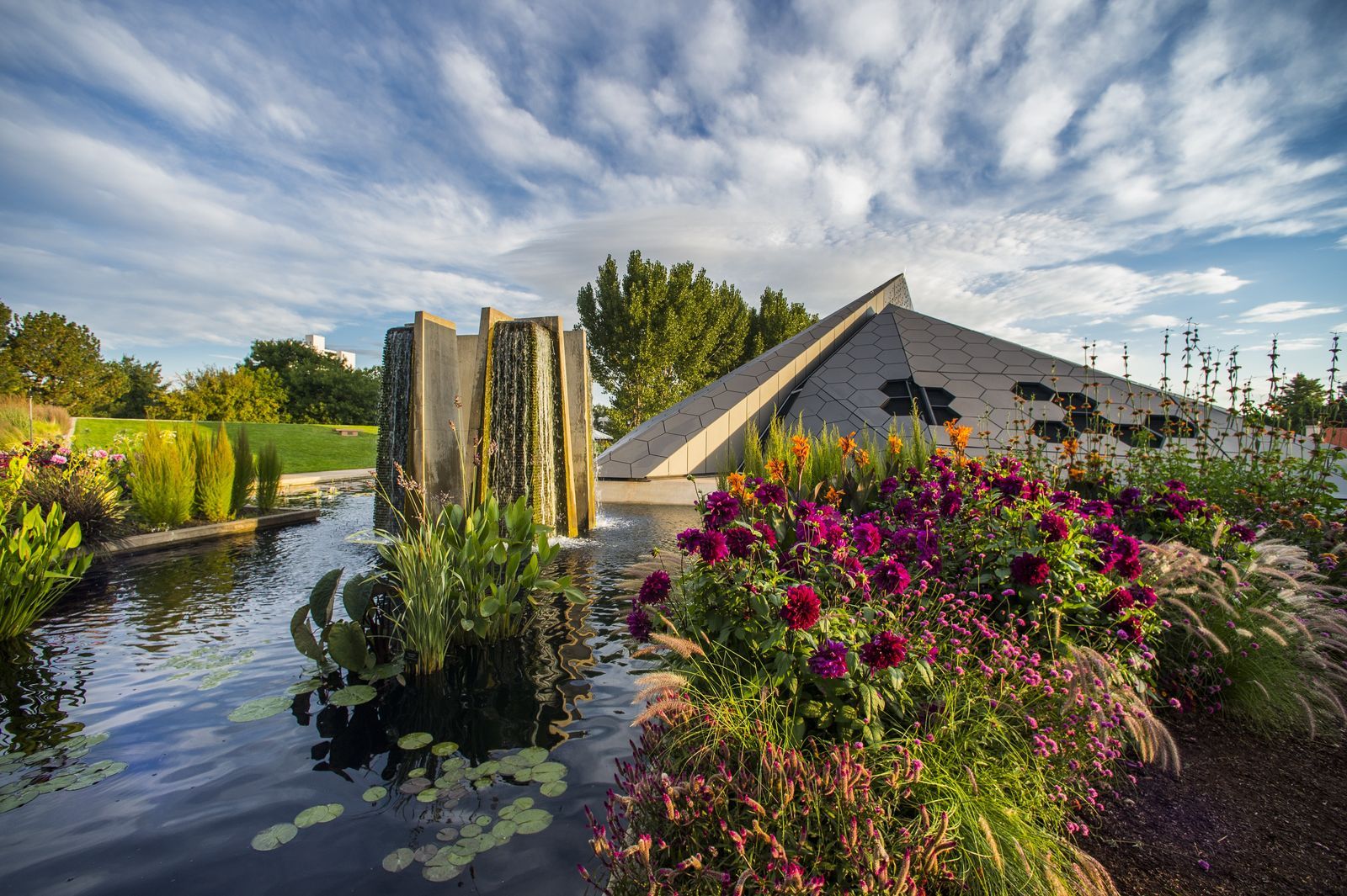 A pond with water lilies and bright pink dahlias in front of a stone fountain sculpture and a pyramid-shaped building under a cloudy sky.
