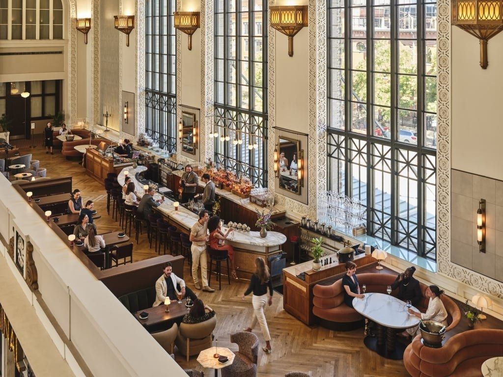A grand hall with tall arched windows, lanterns and a long bar with people seated at tables below.