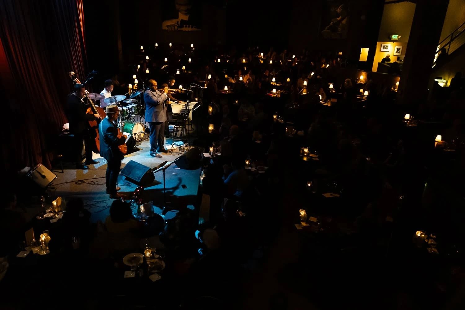 A jazz band on a lit stage in front of a dark dining room full of tables with small lamps.