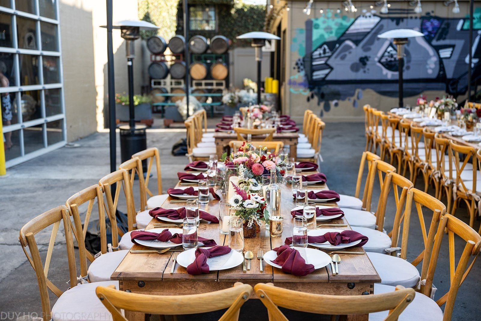 A long banquet table set with plates and flowers under string lights in a courtyard with wine barrels behind.